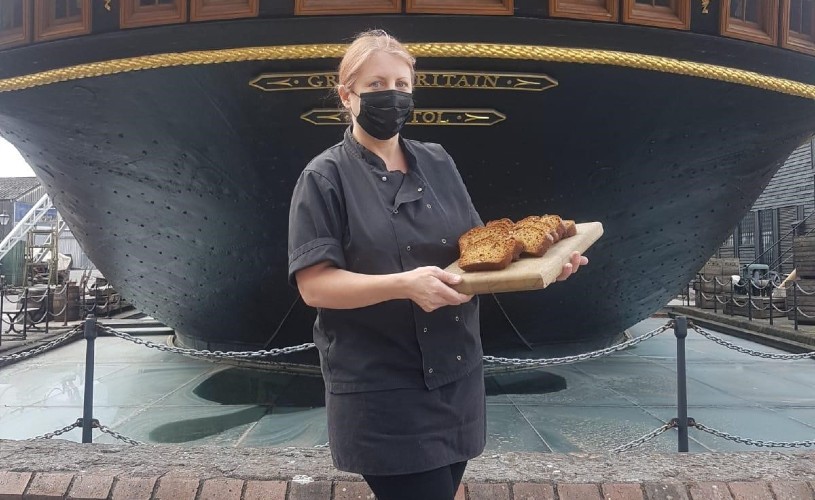 Paula Bishop, baker at Harbourside Kitchen, holding cake in front of Brunel's SS Great Britain